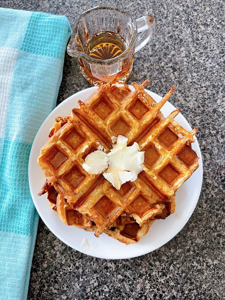 French Toast Waffles topped with butter on a white plate next to a blue and white towel and an empty syrup cup.