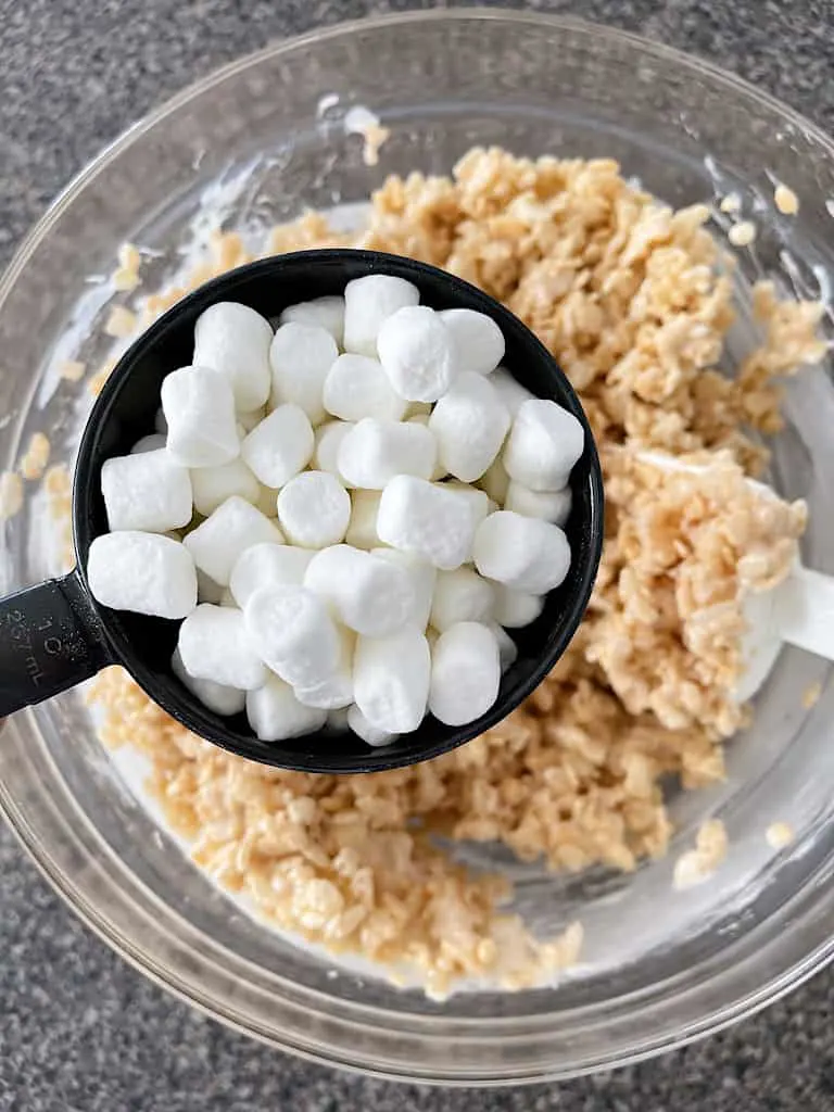 A measuring cup full of mini marshmallows over a bowl of Rice Krispie treats.