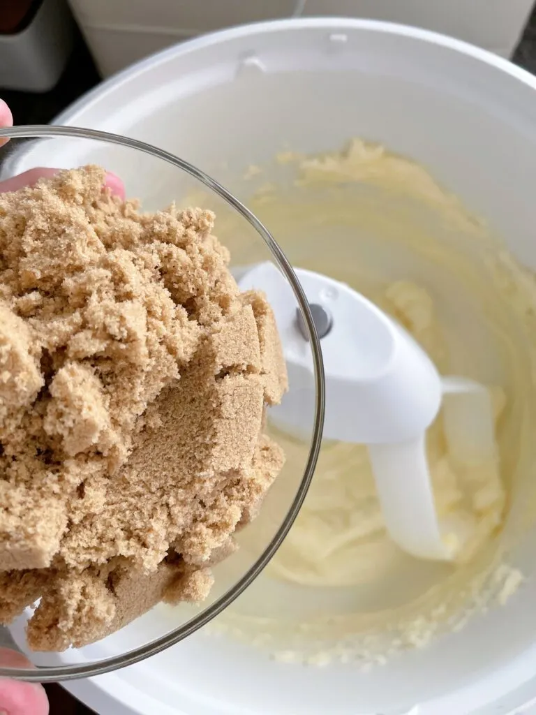 A bowl of brown sugar next to a bowl of cookie dough in a stand mixer.