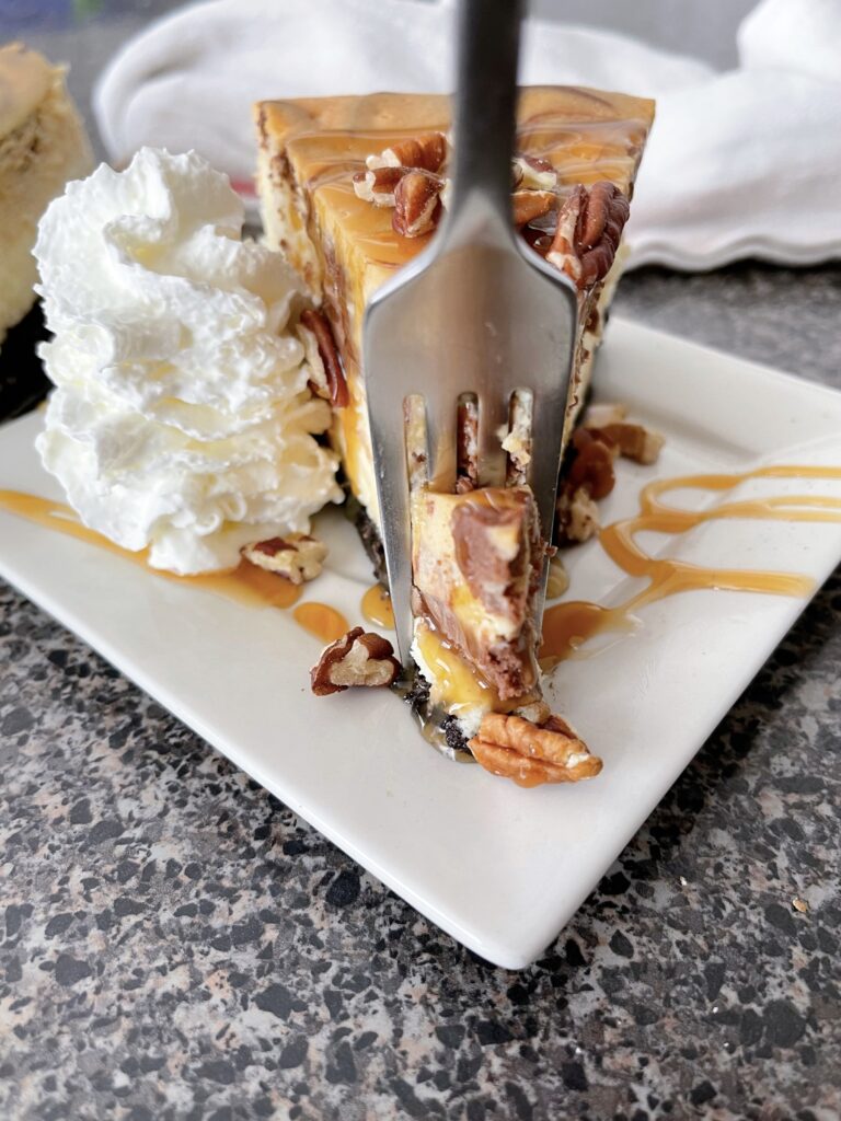 A slice of Cheesecake Factory Caramel Pecan Turtle Cheesecake with whipped cream and caramel sauce.