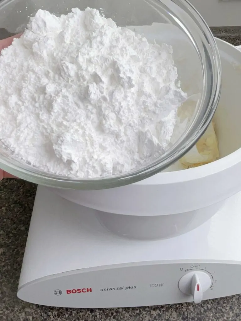 Powdered sugar being added to a mixer with butter to make frosting for cream cheese sugar cookie bars.