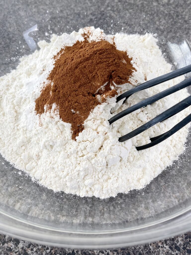 A bowl of flour, baking powder, baking soda and cinnamon with a whisk.