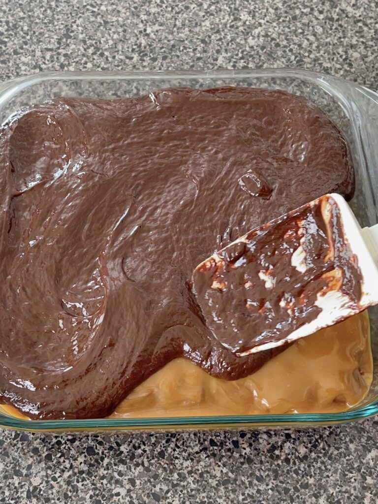 Chocolate ganache being spread over caramel Snickers Brownies to make Choco-Smash Candy Bar Brownies.