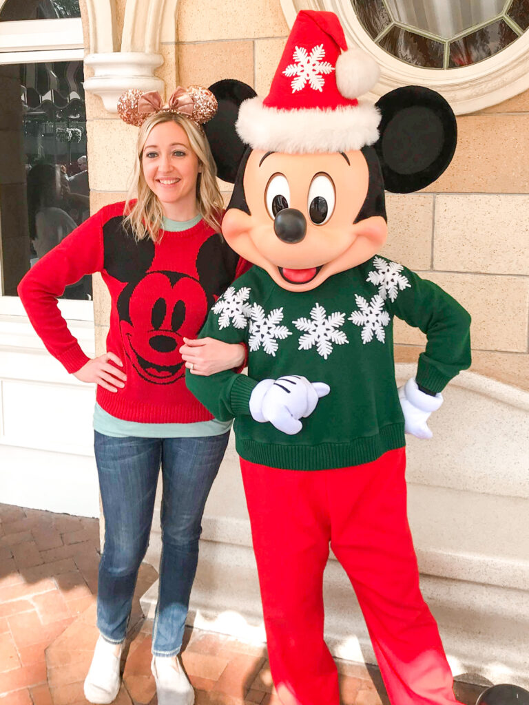 A woman in a Mickey Mouse sweater standing next to Mickey Mouse in a green Christmas sweater.