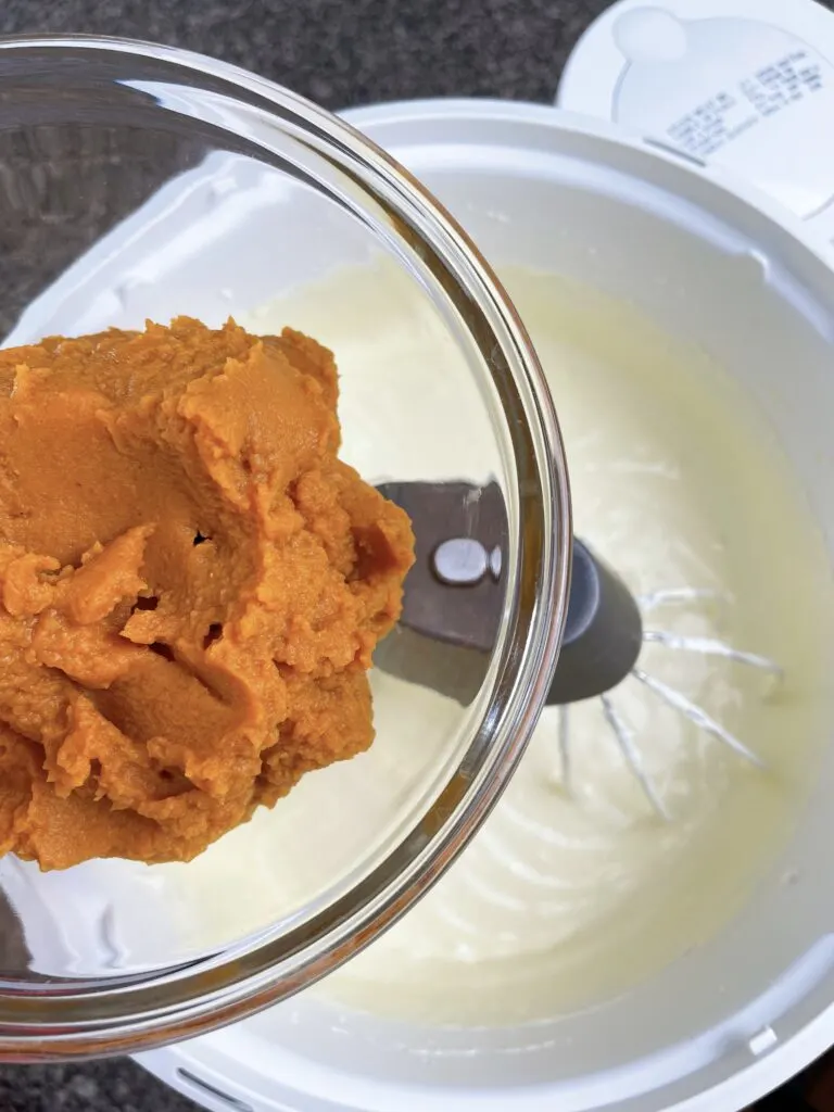A bowl of pumpkin puree over a stand mixer filled with cheesecake batter.
