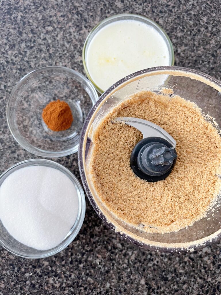 Graham cracker crumbs in a food processor next to bowls of sugar, melted butter, and cinnamon.