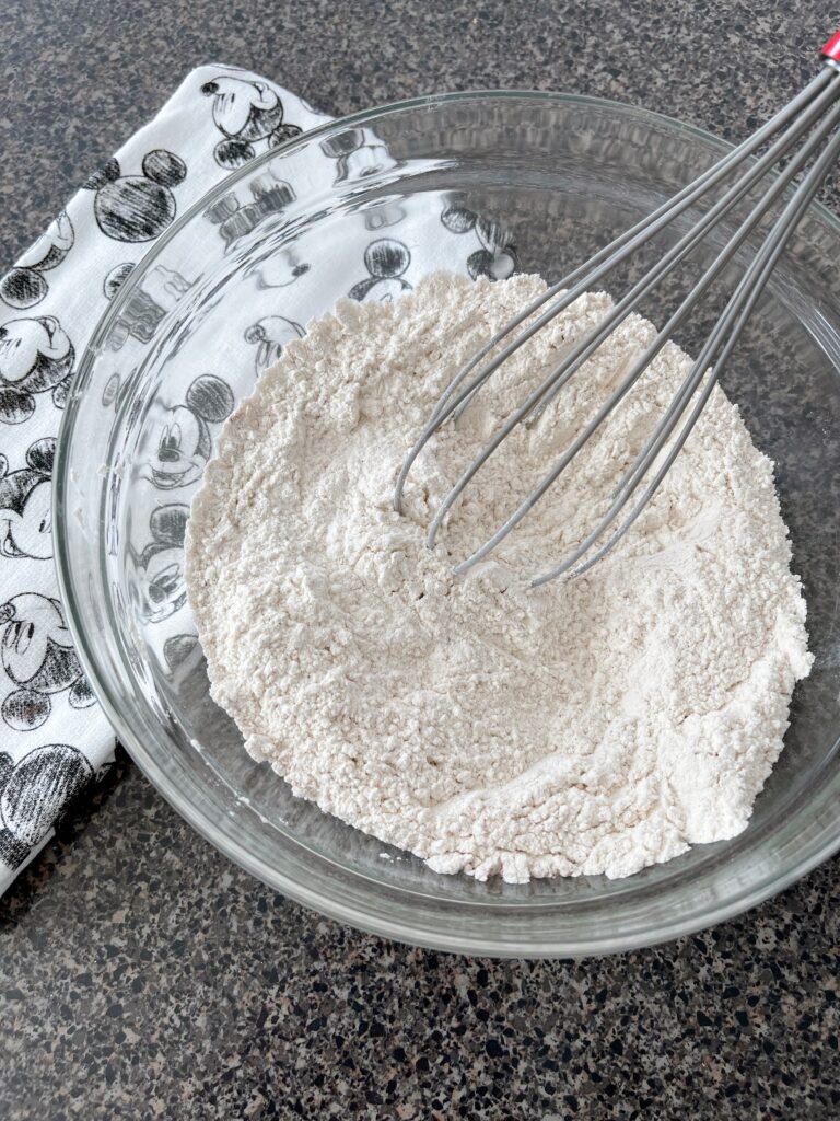 Flour, baking powder, cinnamon, and salt in a mixing bowl with a whisk and a Mickey Mouse towel.