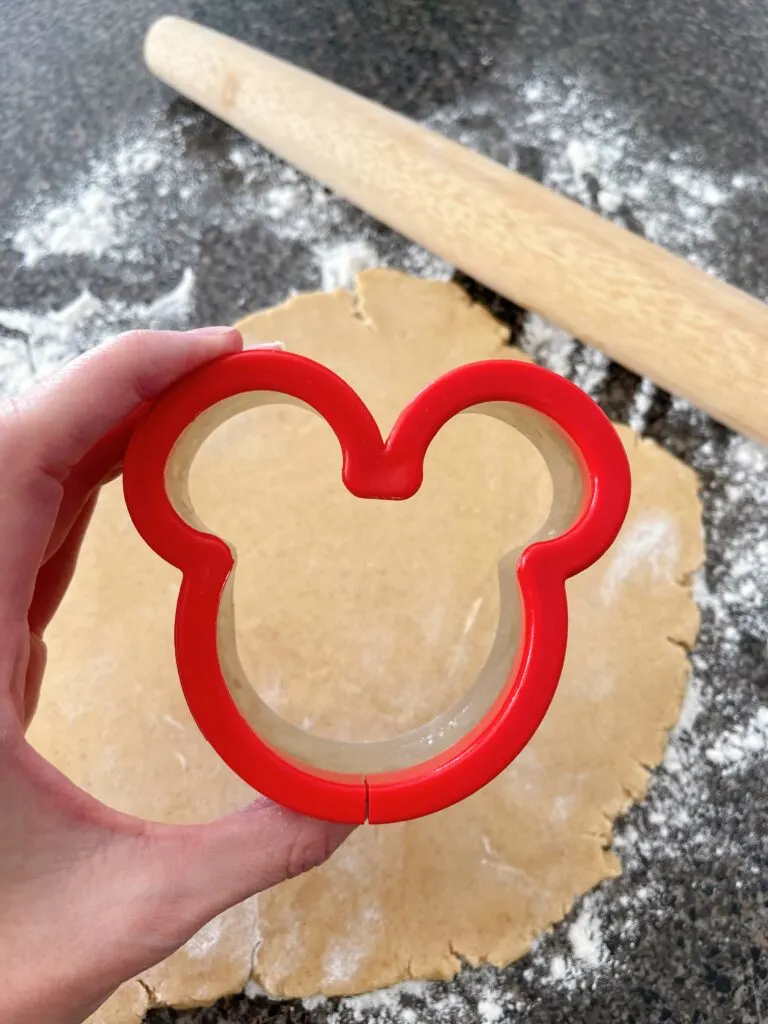 A Mickey Mouse cookie cutter with cookie dough and a rolling pin.
