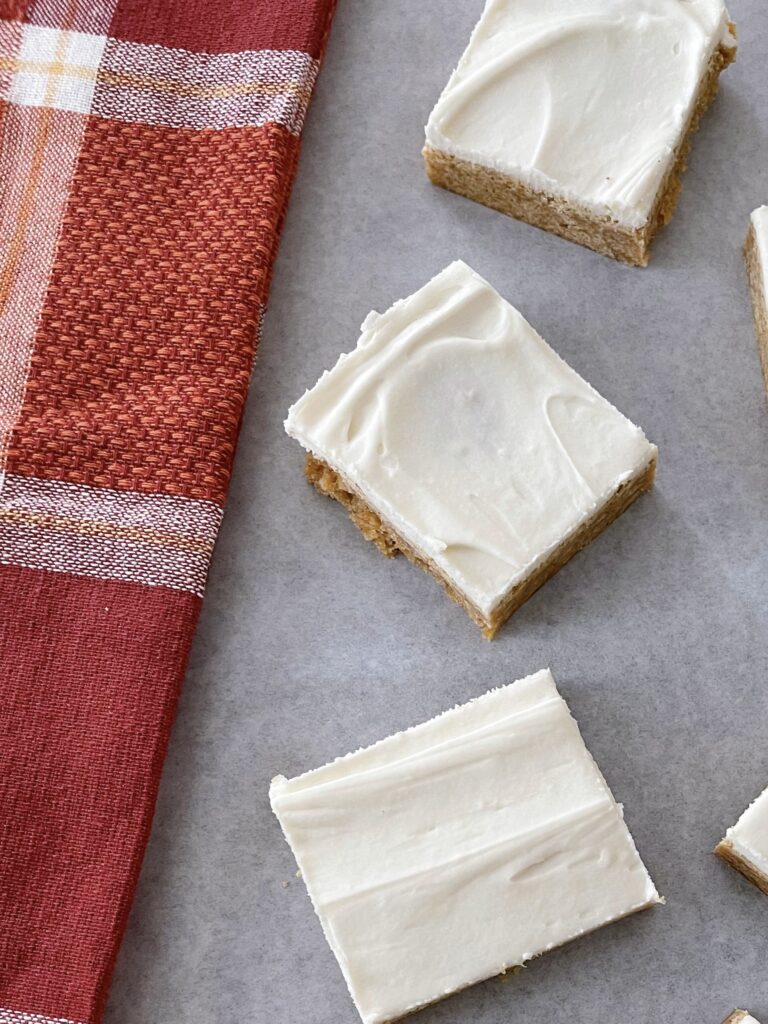 Three pumpkin sugar cookie bars with cream cheese frosting next to a fall colored kitchen towel.