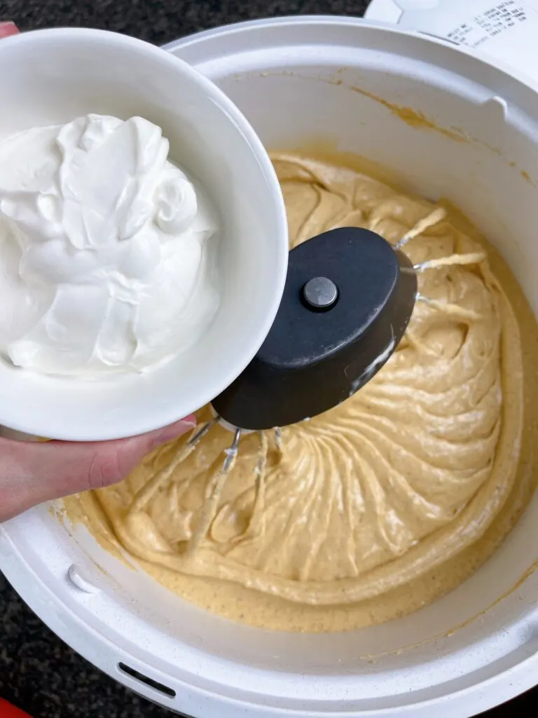 A bowl of sour cream over a stand mixer filled with cheesecake batter.