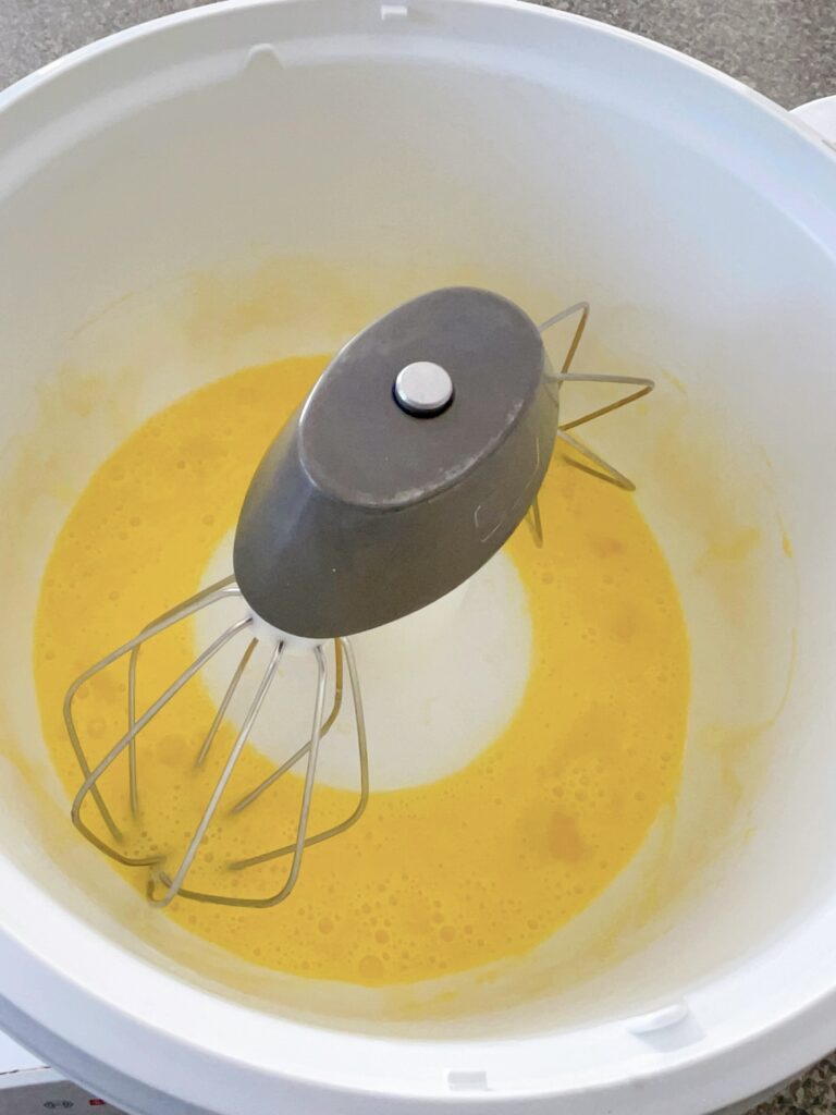 Beaten eggs in the bowl of a stand mixer with a whisk attachment.