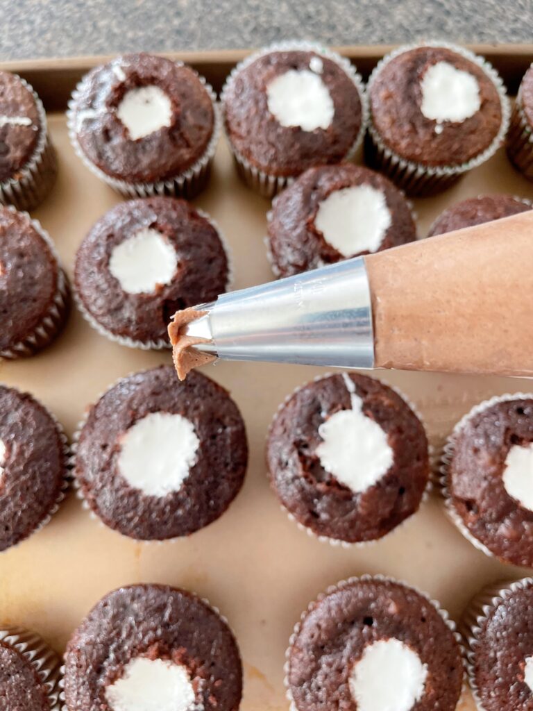 A piping bag with a star tip filled with hot chocolate frosting and cupcakes filled with marshmallow cream.