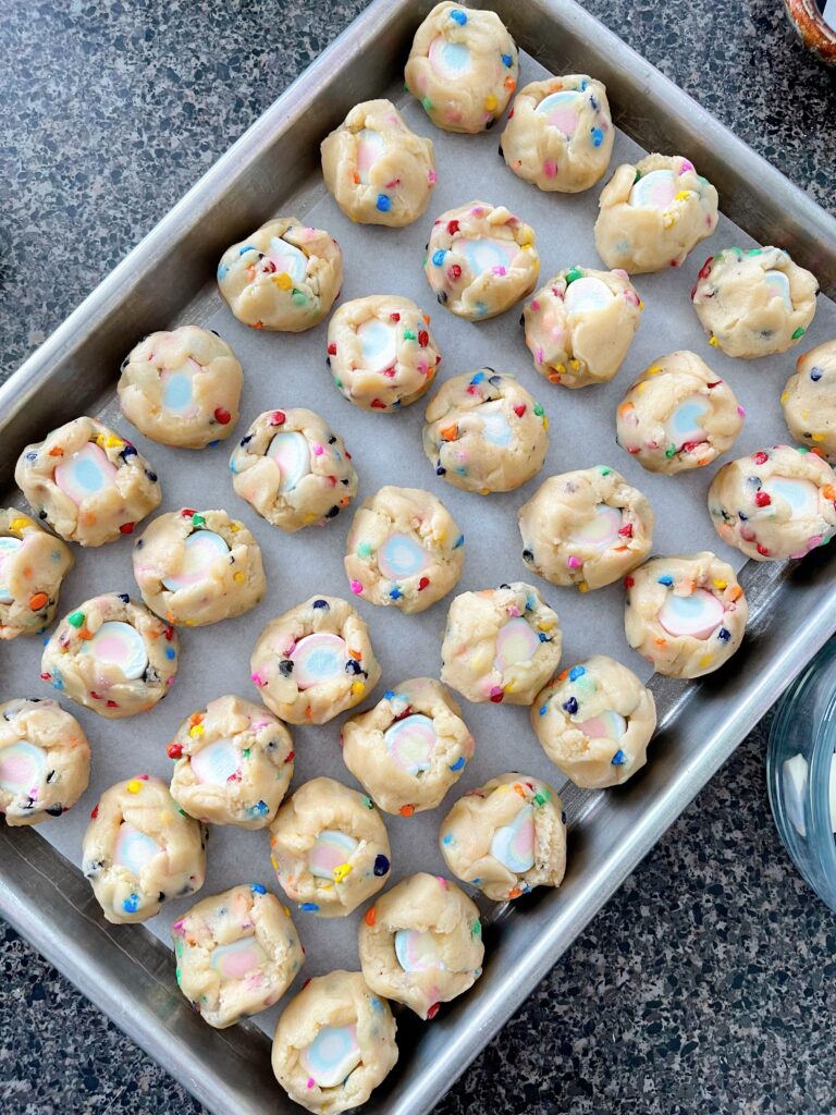 Rainbow chip cookie dough balls stuffed with marshmallows on a baking sheet.