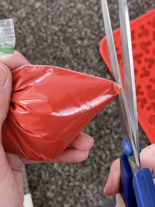 Scissors cutting off the corner of a ziplock bag filled with melted red candy melts.