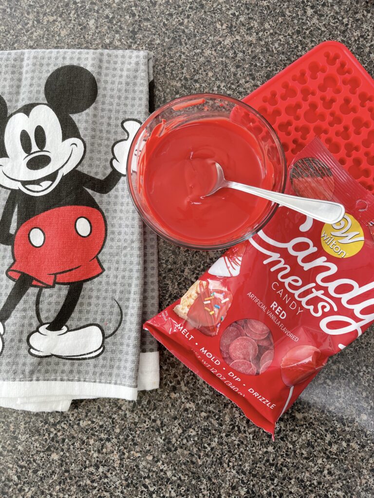 A bowl of melted candy melts with a bag of red candy melts, a Mickey Mouse kitchen towel and a Mickey Mouse silicone mold.