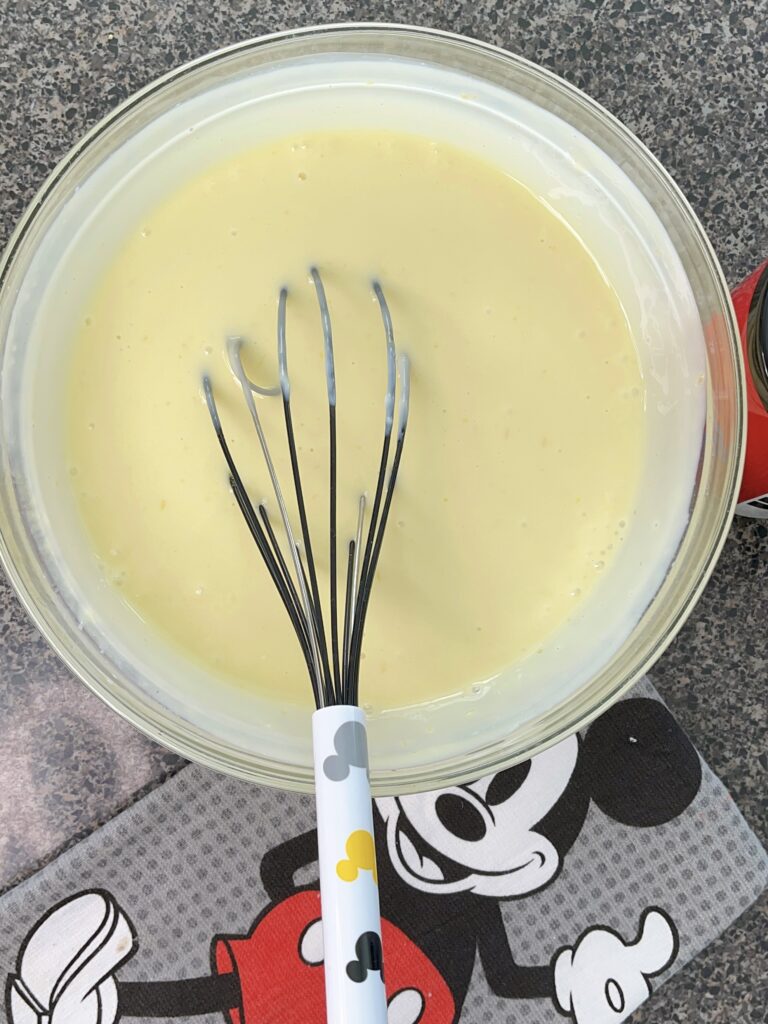Egg yolks mixed with lemon juice and condensed milk in a glass bowl with a Mickey Mouse whisk and kitchen towel.