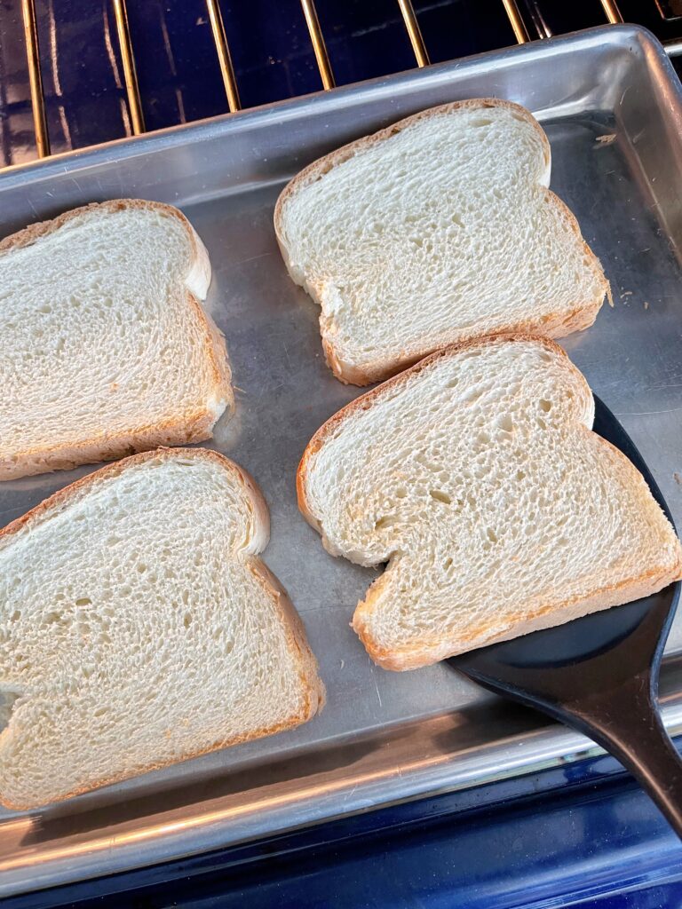 Four slices of bread on a baking sheet with a spatula to flip the bread over.