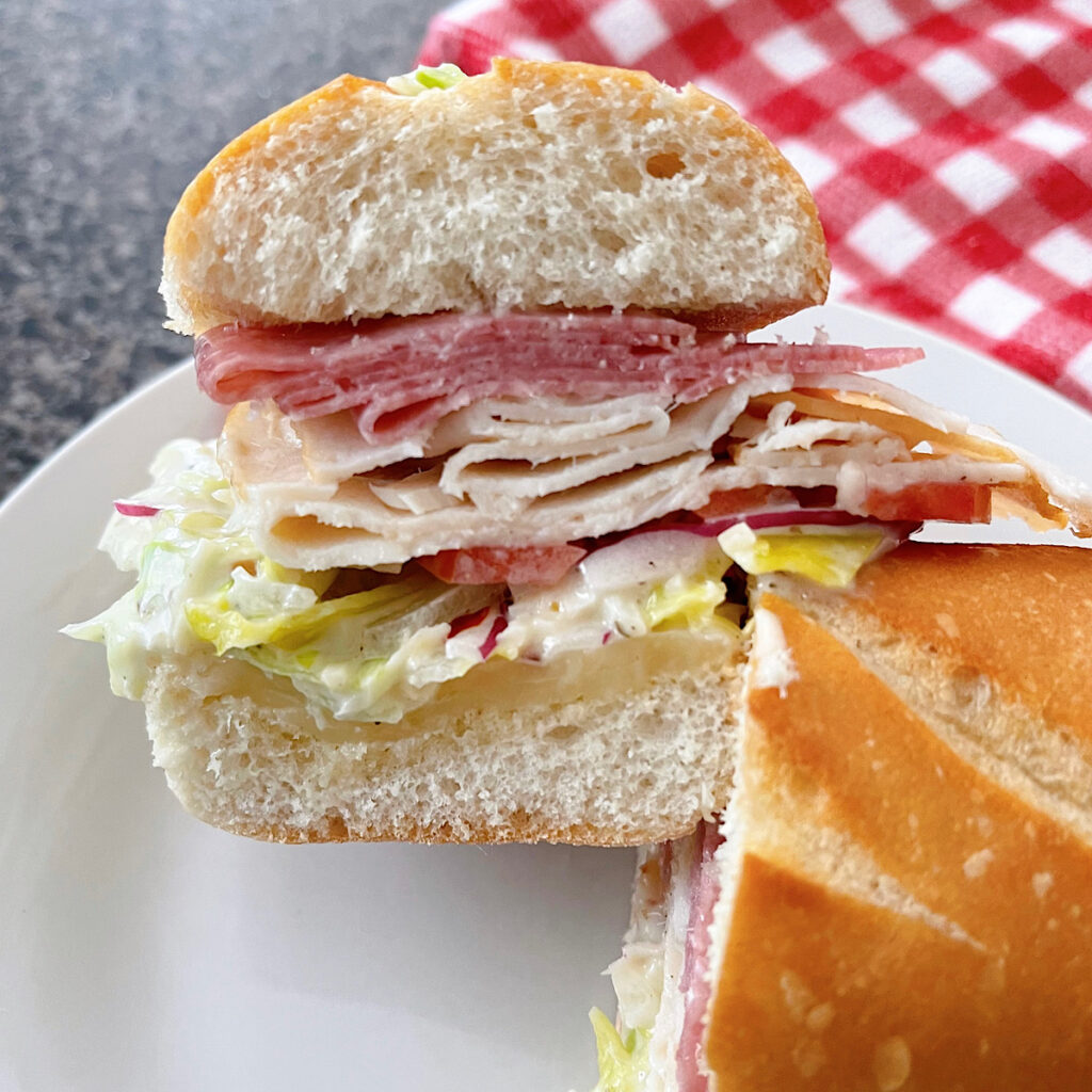 A TikTok grinder sandwich with meat, cheese, and grinder salad on a white plate with a red and white kitchen towel.