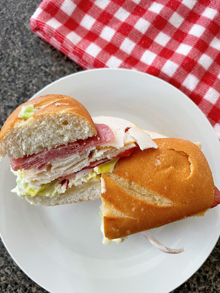 A TikTok grinder sandwich with meat, cheese, and grinder salad on a white plate with a red and white kitchen towel.