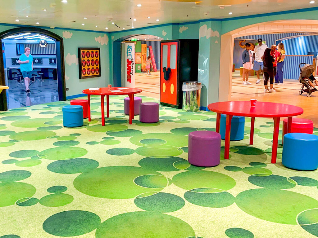 A room with red tables and green Mickey Mouse carpet in the Oceaneer Club on the Disney Magic.