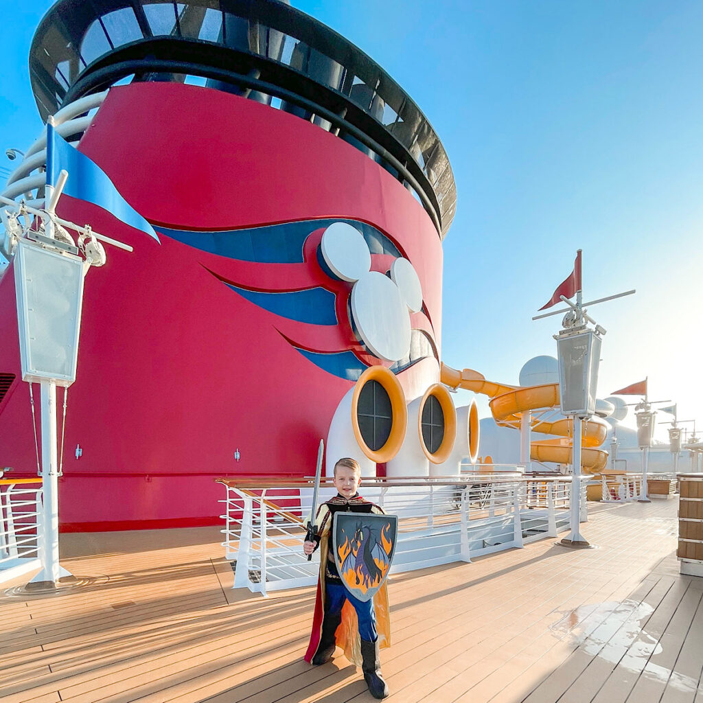 A boy in a prince costume on the top Deck of the Disney Magic in front of the funnel with a which Mickey Mouse head.