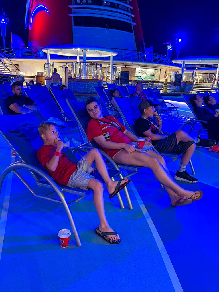Three kids sitting in lounge chairs watching a movie on a giant screen on the Disney Magic.