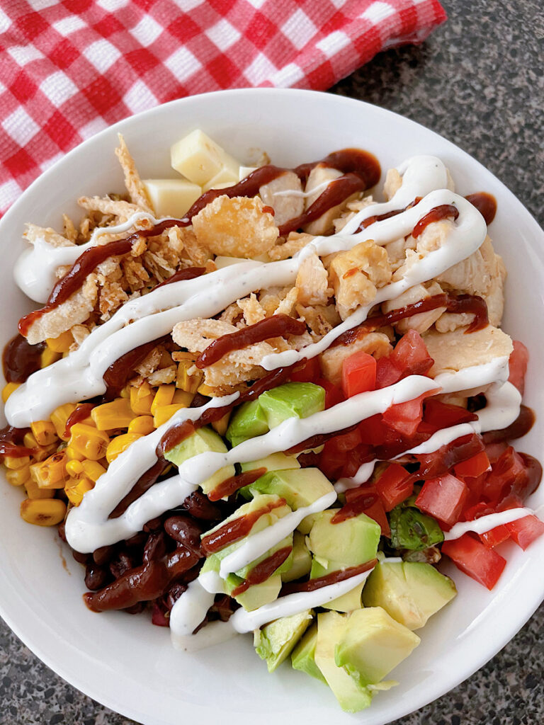 A barbecue chicken salad in a white bowl with black beans, corn, avocado, tomatoes, pepper jack, corn, chicken, and fried onions drizzled with bbq sauce and ranch dressing.