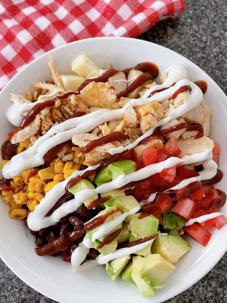 A barbecue chicken salad in a white bowl with black beans, corn, avocado, tomatoes, pepper jack, corn, chicken, and fried onions drizzled with bbq sauce and ranch dressing.