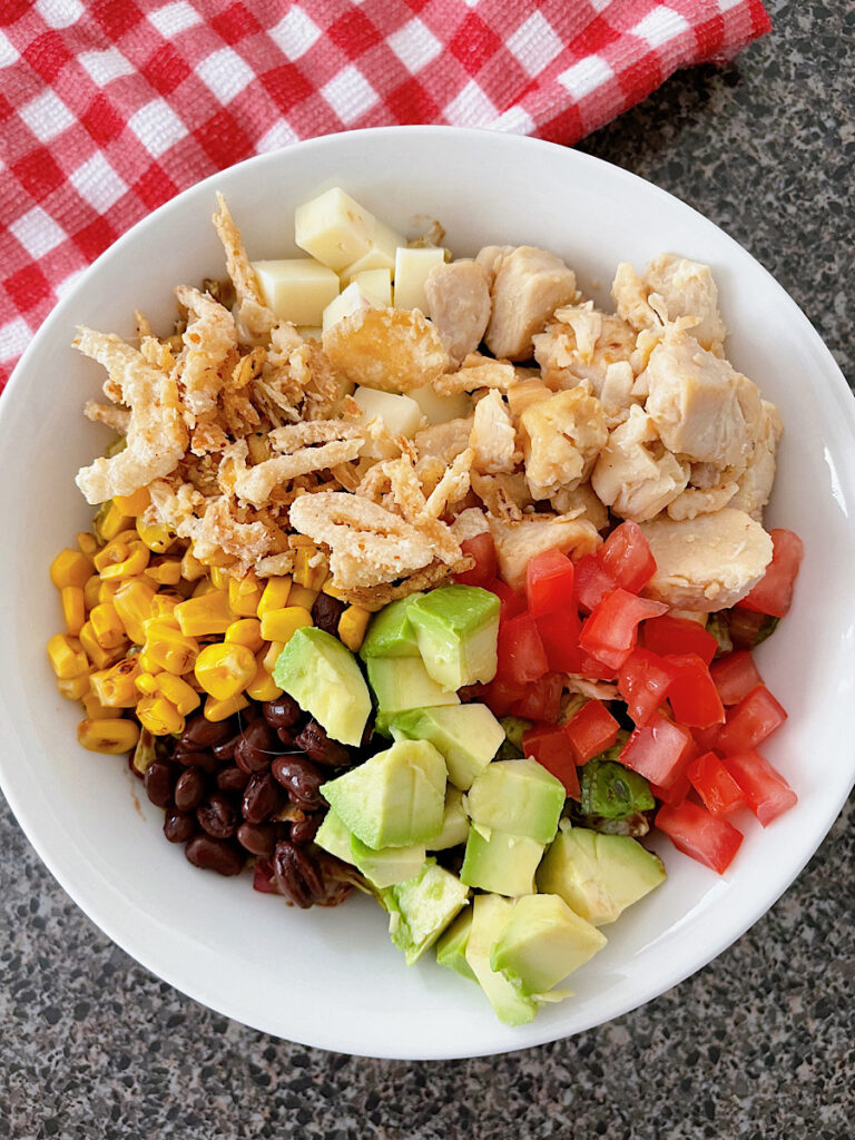 A barbecue chicken salad in a white bowl with black beans, corn, avocado, tomatoes, pepper jack, corn, chicken, and fried onions.
