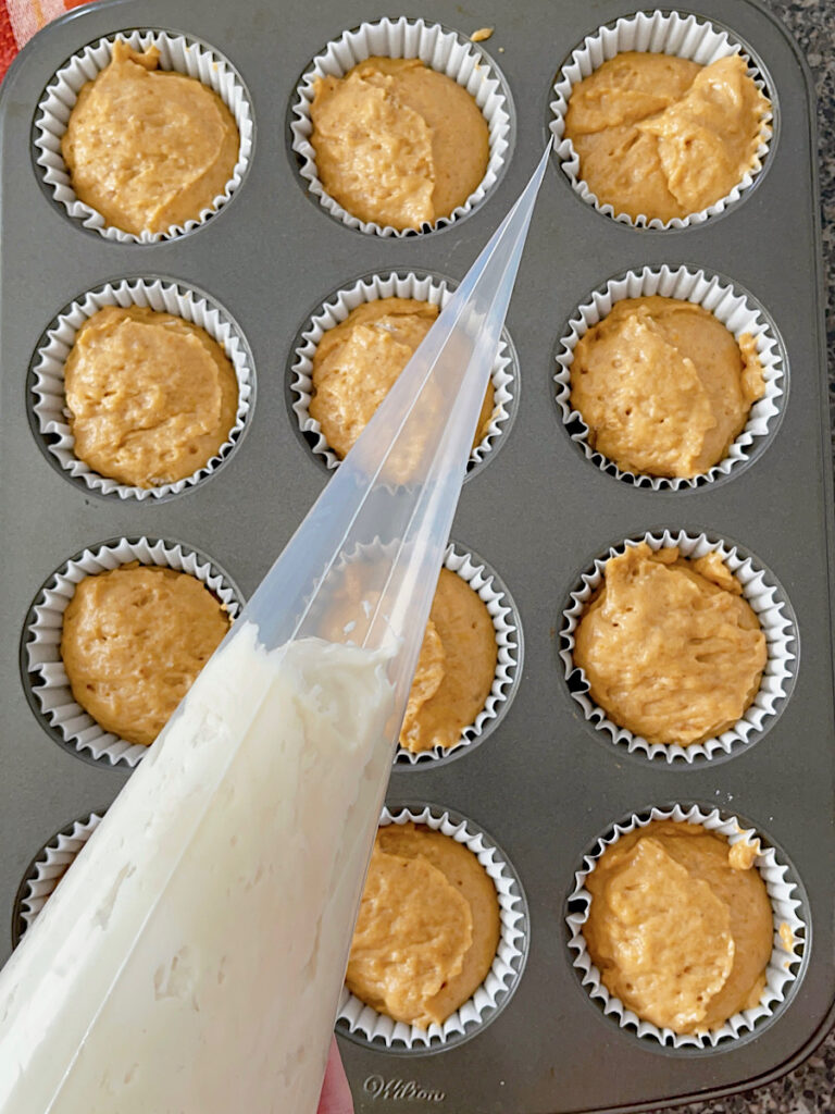 A piping bag filled with cream cheese over a pan of pumpkin muffins.