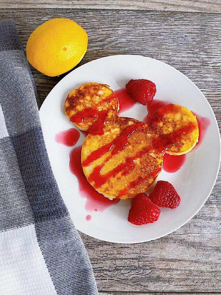 Mickey Mouse shaped Lemon Ricotta Pancakes on a plate with raspberries and raspberry syrup.