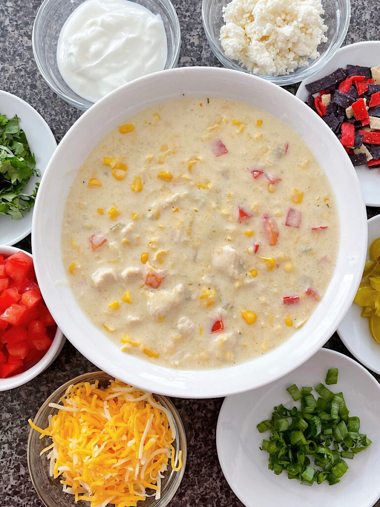 A bowl of corn chowder surrounded by small bowls of assorted toppings.