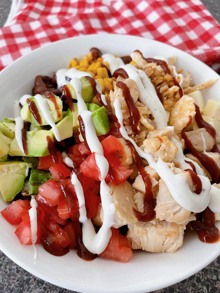 A barbecue chicken salad in a white bowl with black beans, corn, avocado, tomatoes, pepper jack, corn, chicken, and fried onions drizzled with bbq sauce and ranch dressing.