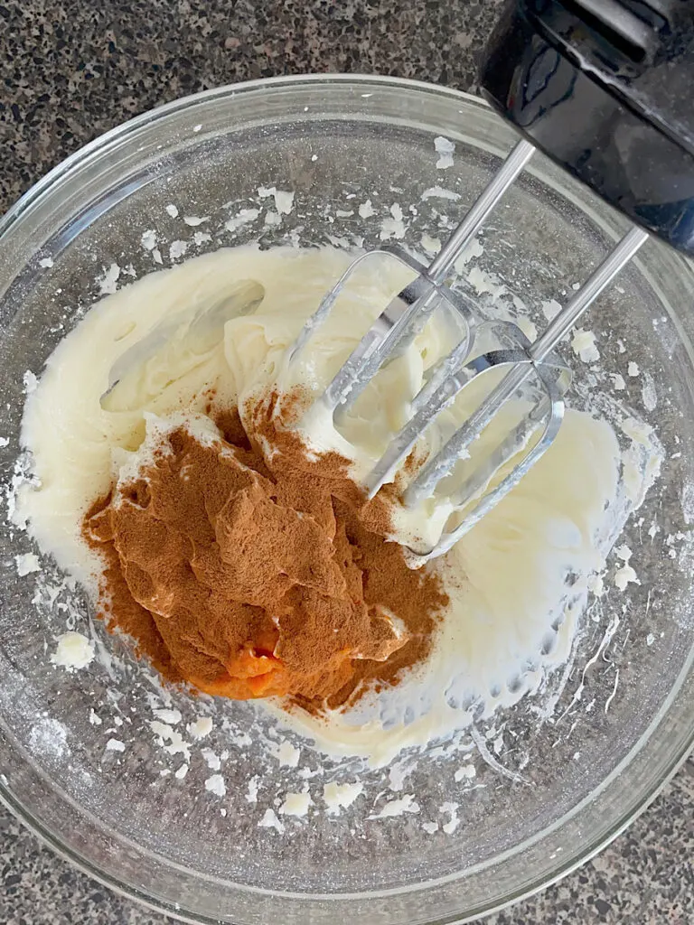 Pumpkin pie spice, cream cheese, and pumpkin puree in a mixing bowl with a hand mixer.