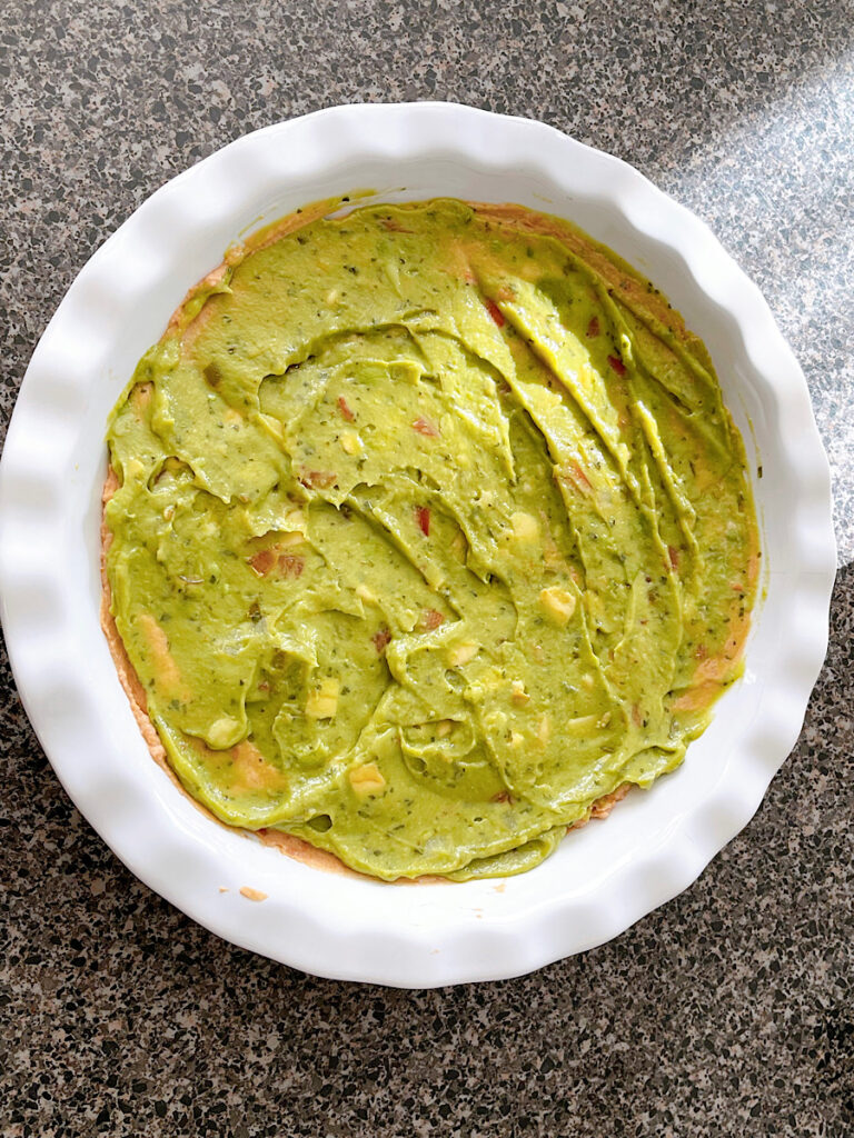 Guacamole spread over beans in a white pie dish for seven layer dip.
