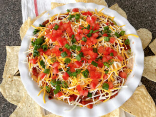 Seven layer dip with beans, cheese, sour cream, guacamole, tomatoes, and onions in a white dish.