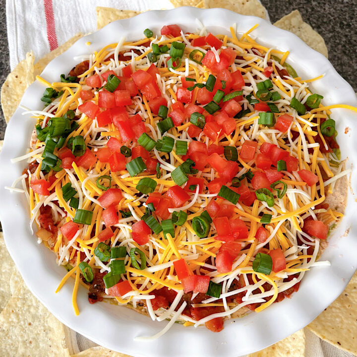 Seven layer dip with beans, cheese, sour cream, guacamole, tomatoes, and onions in a white dish.