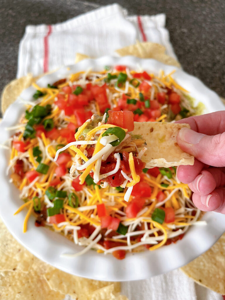 Seven layer dip with beans, cheese, sour cream, guacamole, tomatoes, and onions in a white dish.