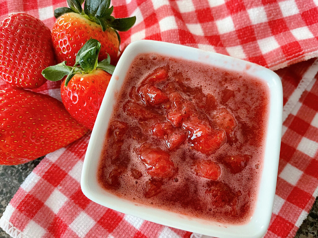 A dish of strawberry compote next to fresh strawberries on a red and white towel.