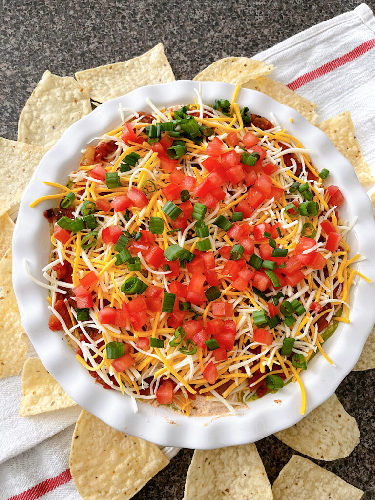 Seven layer dip with beans, cheese, sour cream, guacamole, tomatoes, and onions in a white dish.