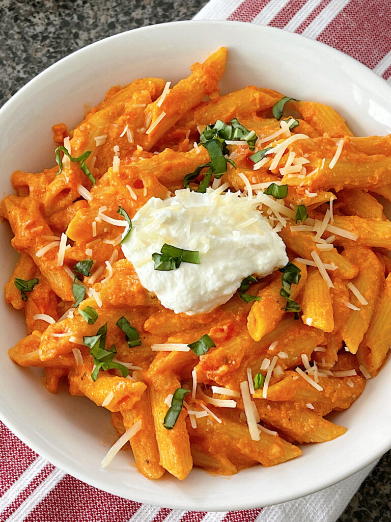 A bowl of homemade Cheesecake Factory Four Cheese Pasta topped with ricotta cheese and chopped fresh basil.