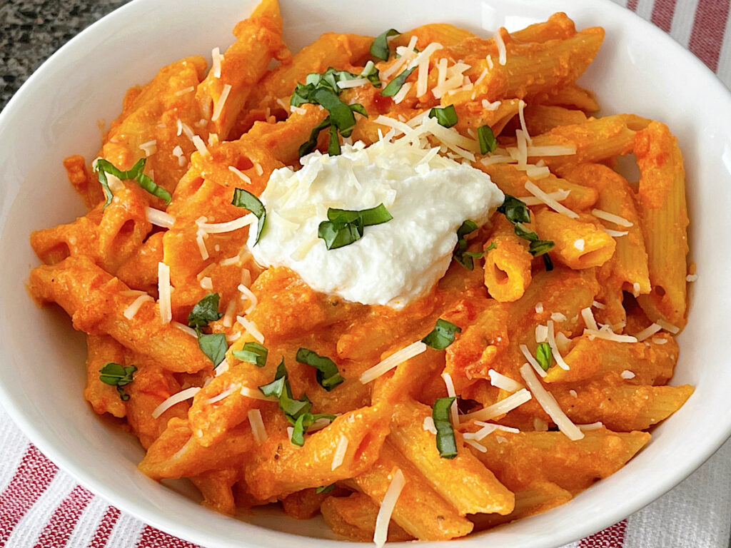 A bowl of homemade Cheesecake Factory Four Cheese Pasta topped with ricotta cheese and chopped fresh basil.