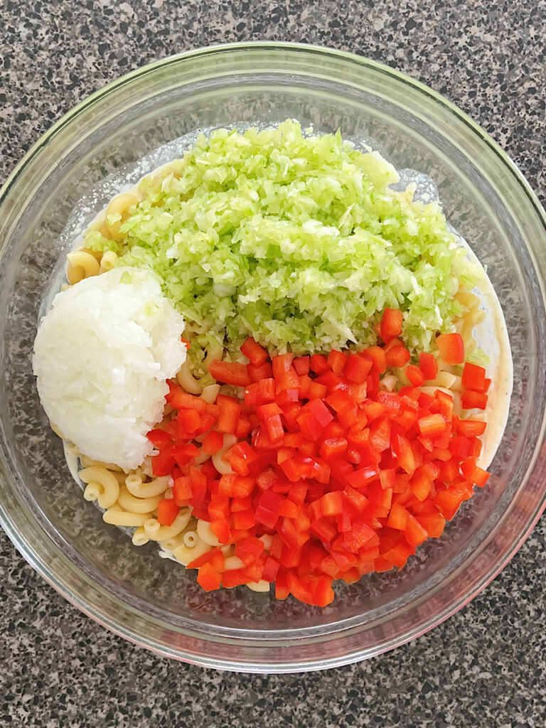 Celery, macaroni, red bell pepper, and onion in a bowl with macaroni salad dressing.