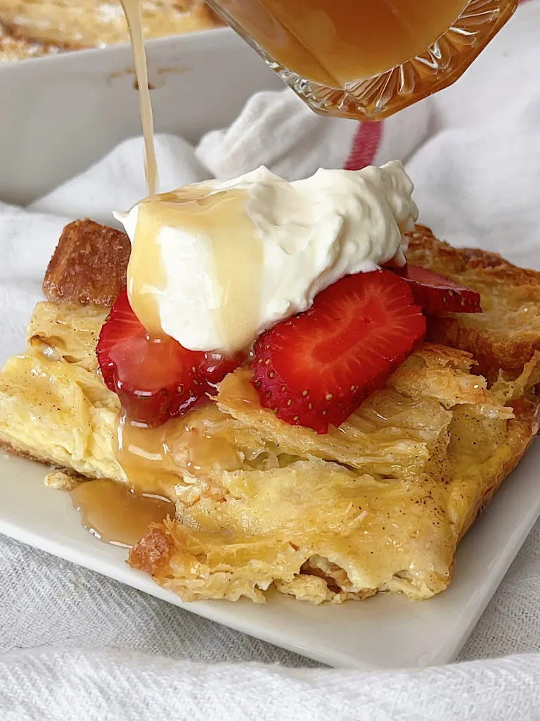 A slice of a croissant French toast overnight casserole topped with whipped cream, caramel syrup and strawberries.