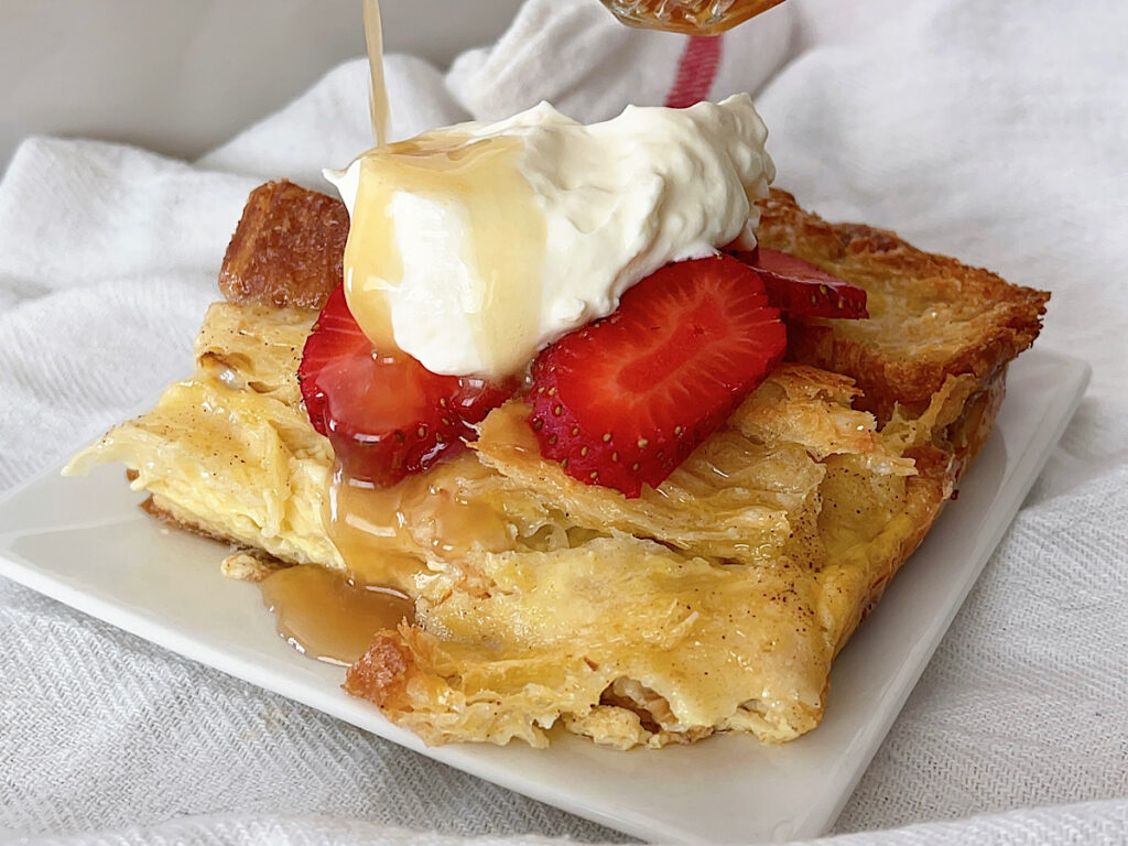 A slice of a croissant French Toast casserole topped with strawberries, whipped cream, and caramel syrup.