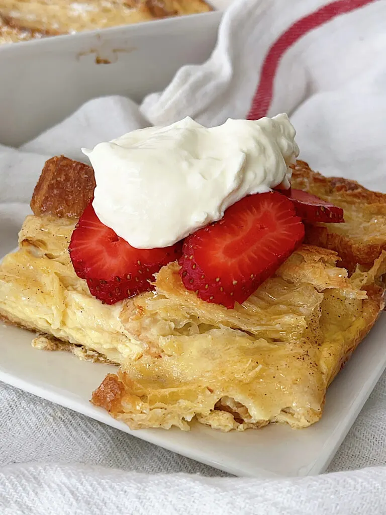 A slice of a croissant French toast casserole topped with whipped cream and strawberries.