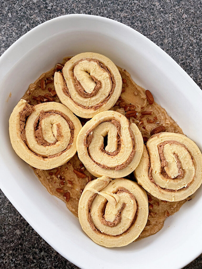 Pillsbury cinnamon rolls on top of brown sugar and pecans in a white baking dish.