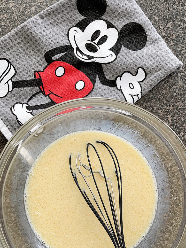 A bowl of wet ingredients to make Churro Mickey Waffles and a Mickey Mouse towel.