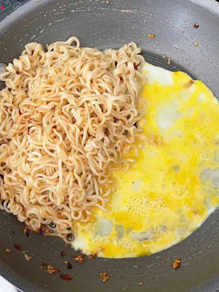 Ramen noodles in a spicy homemade sauce next to eggs cooking in a skillet.