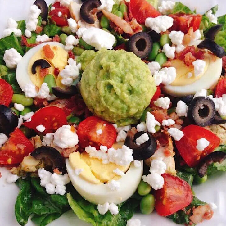 A bowl of lettuce topped with hard boiled egg, black olives, tomatoes, feta cheese, bacon, edamame, and mashed avocado.